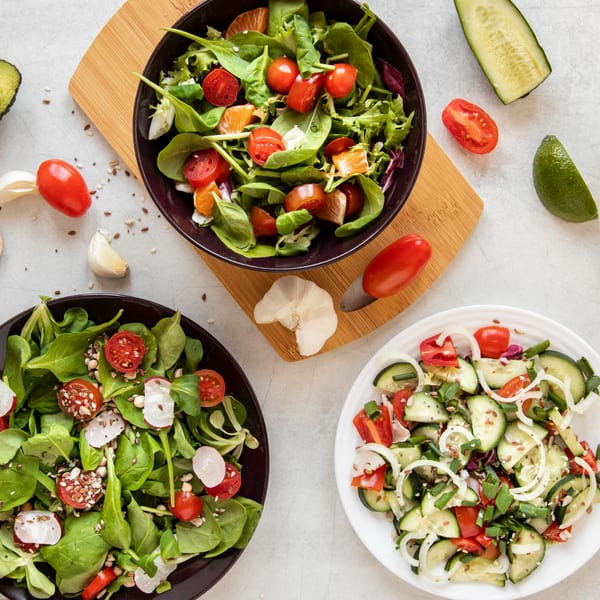 Three bowls of fresh salads with various vegetables, seeds, and fruits on a light background.