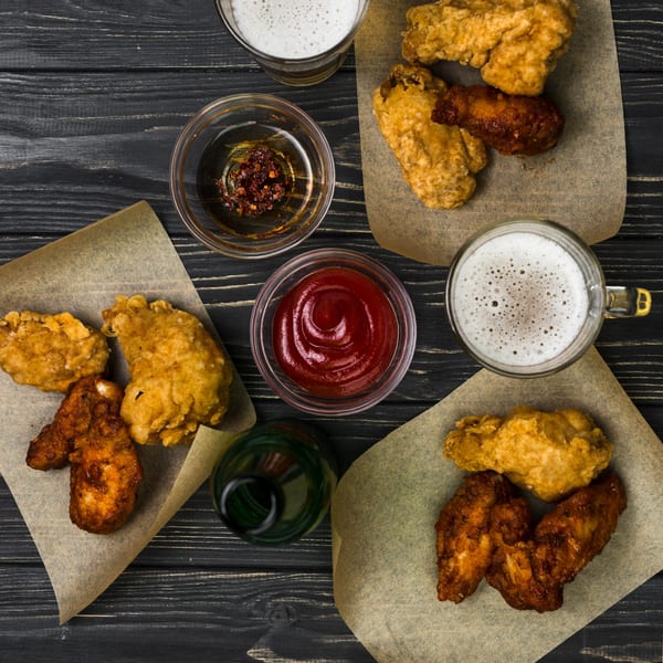 Top-down view of fried chicken wings, beer, ketchup, and chili oil on a dark wooden table.
