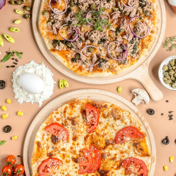 Two pizzas, one with tuna and red onion, the other with tomatoes, surrounded by ingredients.