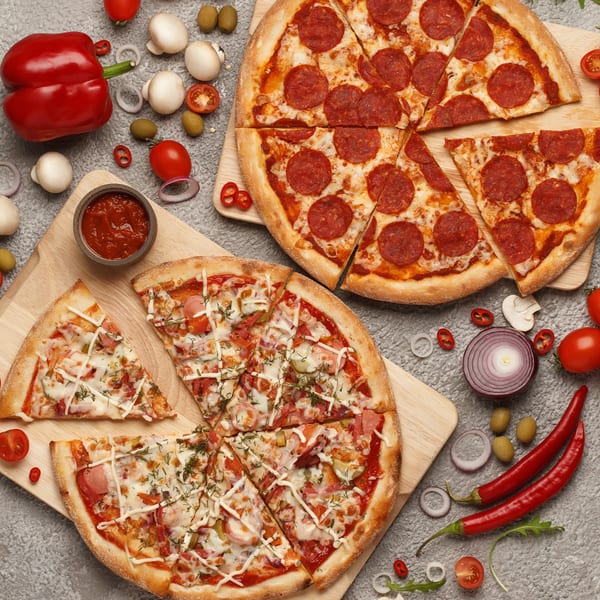 Two Milton New York pizzas, one pepperoni and one with various toppings, surrounded by fresh ingredients.
